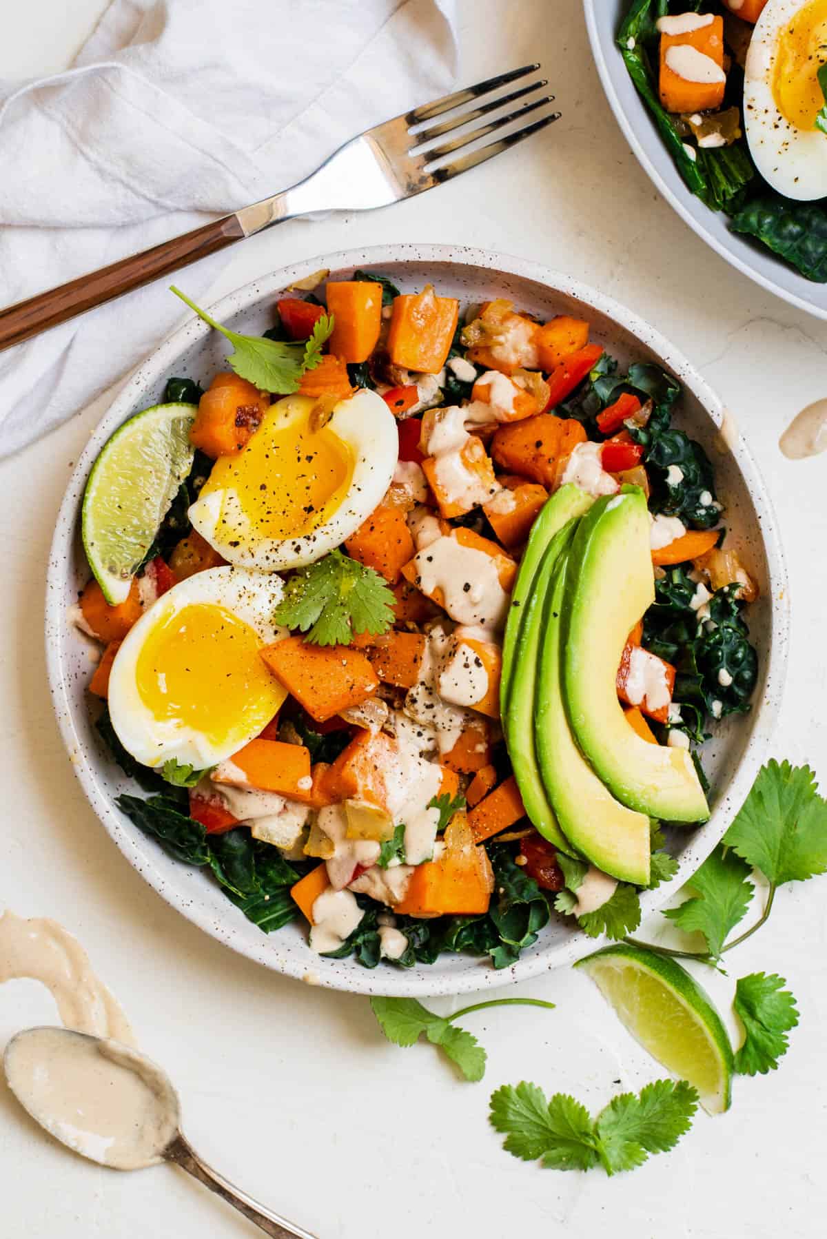 Sweet potato breakfast bowl layered over kale and topped with sliced avocado, boiled eggs, cilantro, lime wedge and a drizzle of tahini.