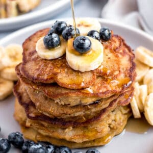A stack of banana oat pancakes on a plate with sliced bananas and fresh blueberries on top. Maple syrup is drizzled on top.