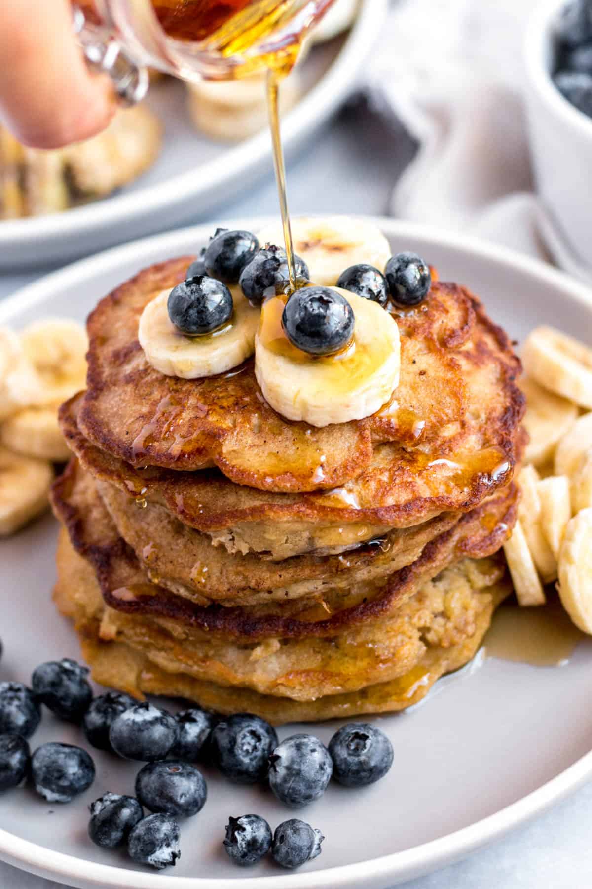 A stack of banana oat pancakes on a plate with sliced bananas and fresh blueberries on top. Maple syrup is drizzled on top.