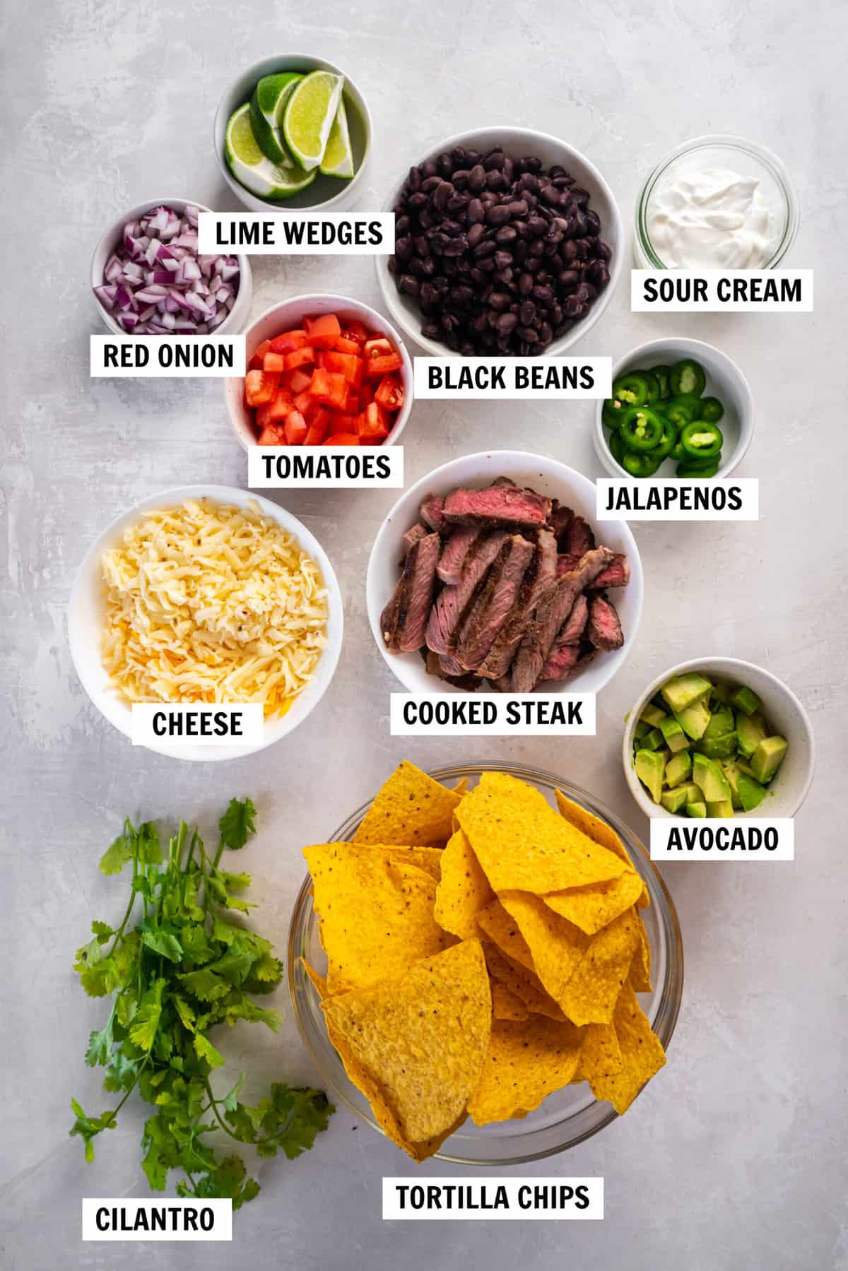 All of the ingredients for steak nachos on a white tabletop including cooked steak, tortilla chips, cheese, onion, tomato, jalapenos, avocado, sour cream and cilantro.