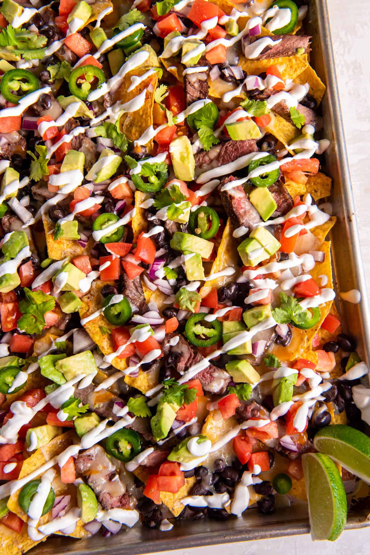 Loaded steak nachos on a sheet pan with all of the toppings including steak, tomatoes, cheese, avocado, jalapenos and sour cream. 