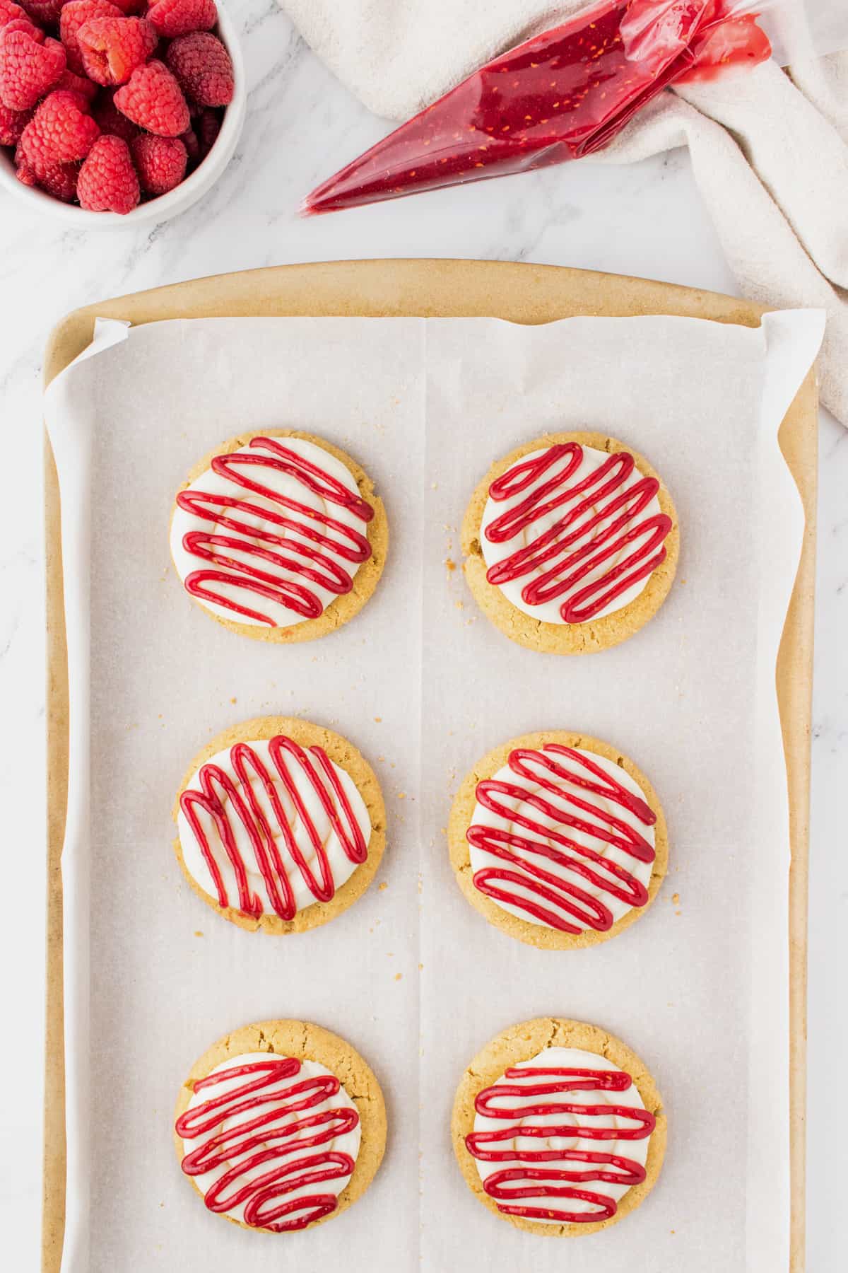 Raspberry cheesecake cookies on a baking sheet after the raspberry preserves are drizzled on top of the cream cheese frosting.