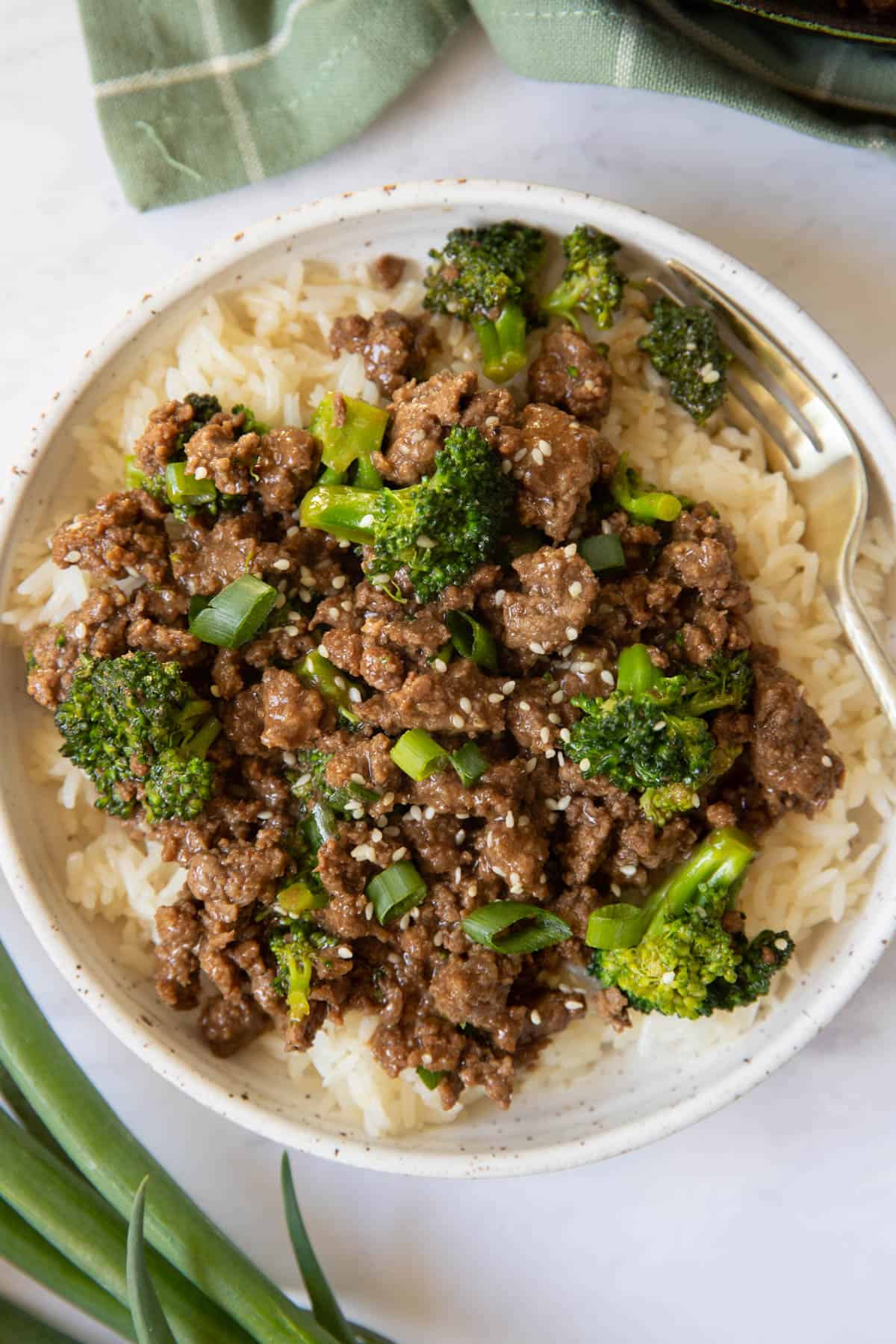 Ground beef and broccoli served on a white plate with a gold fork. Sesame seeds and sliced green onions are sprinkled on top for garnish.