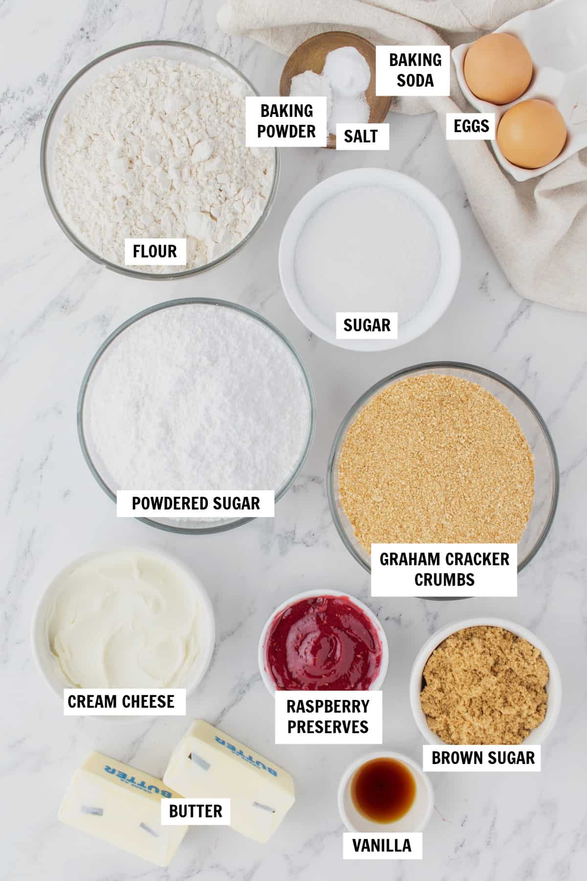 All of the ingredients you need for Raspberry Cheesecake Cookies sitting on a white countertop including butter, sugar, eggs, vanilla, flour, graham cracker crumbs, salt, baking powder, baking soda, cream cheese and raspberry preserves.