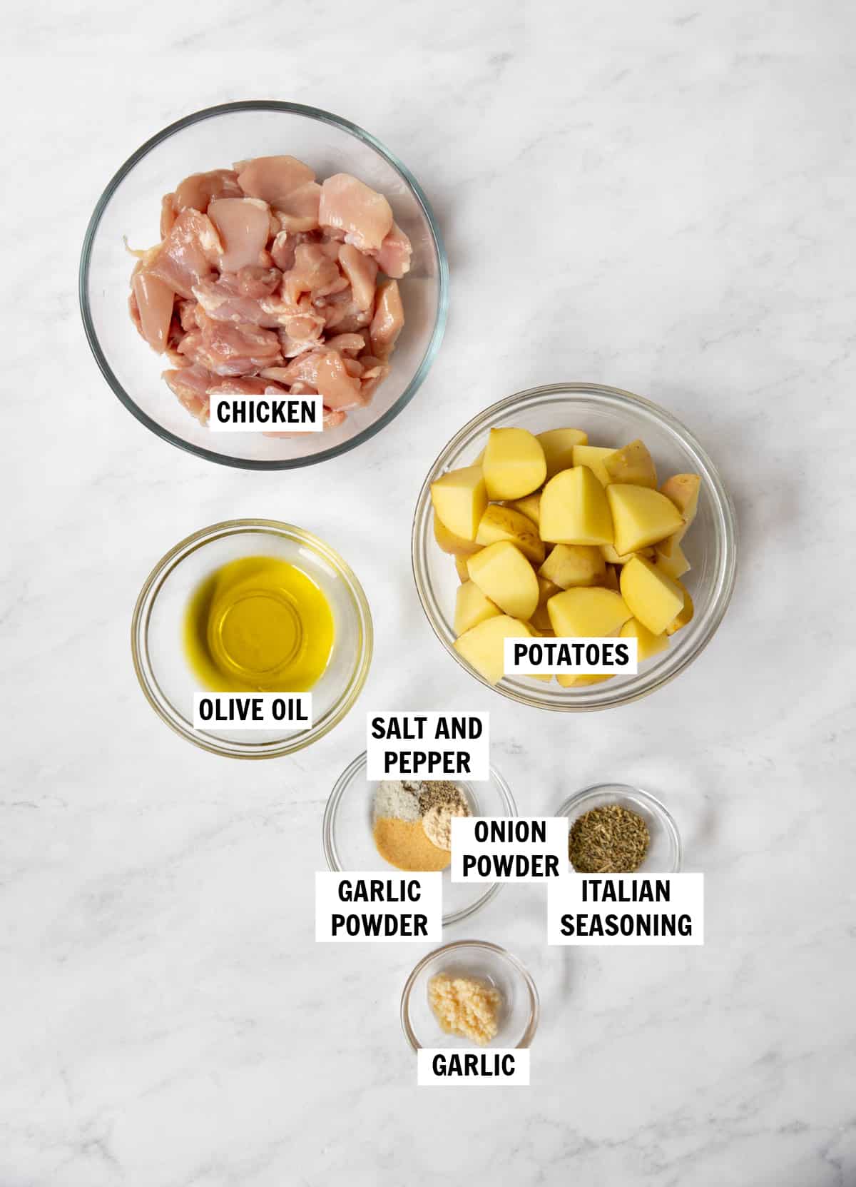 All of the ingredients for chicken and potatoes on a white countertop in bowls including chicken thighs, yellow potatoes, olive oil, garlic powder, onion powder, italian seasoning, garlic and salt and pepper.
