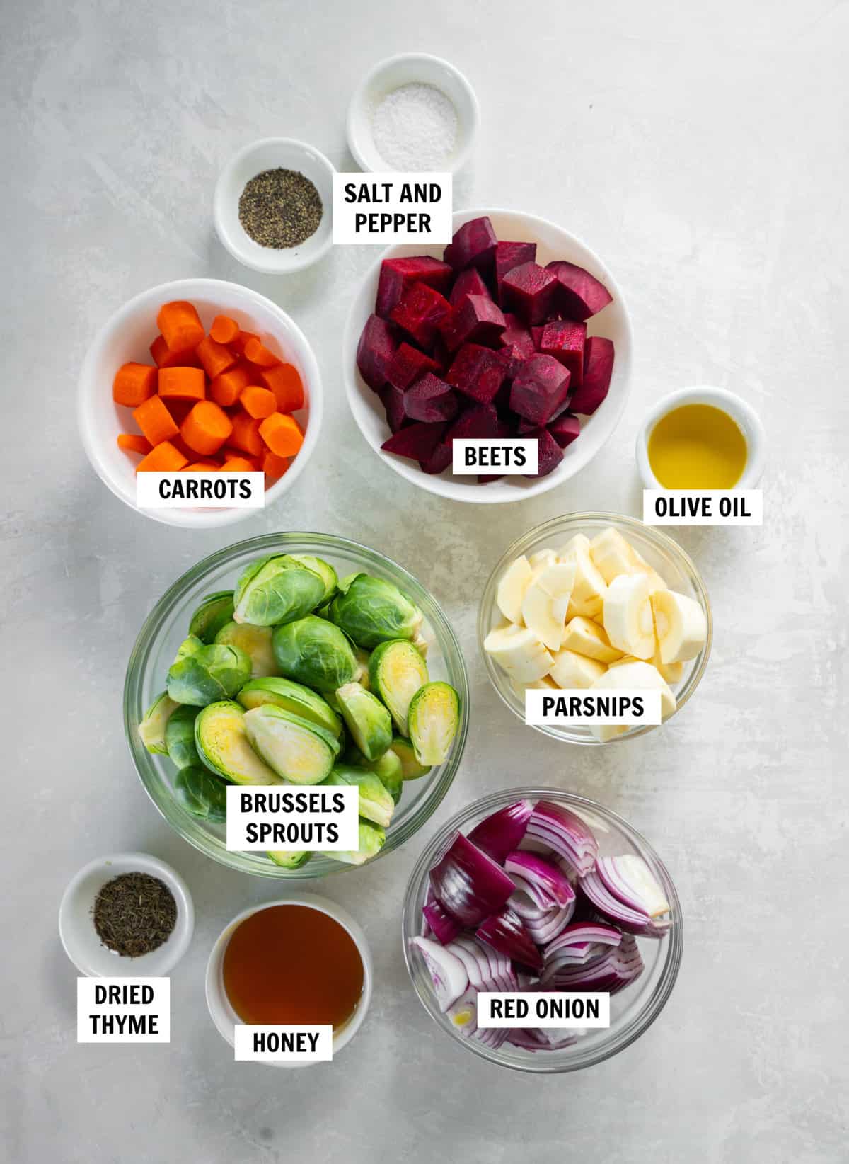 All of the ingredients for honey roasted vegetables on a white countertop in bowls including beets, brussels sprouts, carrots, parsnips, red onion, honey, olive oil, thyme, salt and pepper.