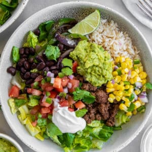 A white bowl filled with rice, cooked ground beef, black beans, corn salsa, diced tomatoes, guacamole and sour cream to make a beef burrito bowl.