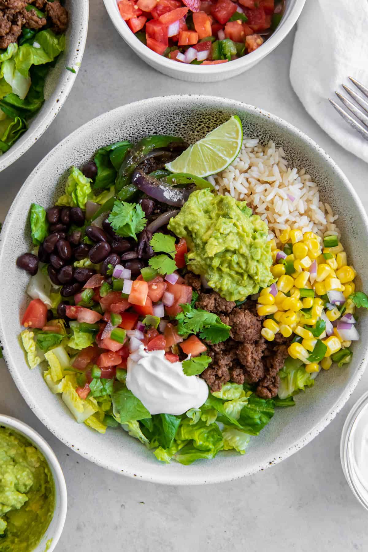 A white bowl filled with rice, cooked ground beef, black beans, corn salsa, diced tomatoes, guacamole and sour cream to make a beef burrito bowl. 