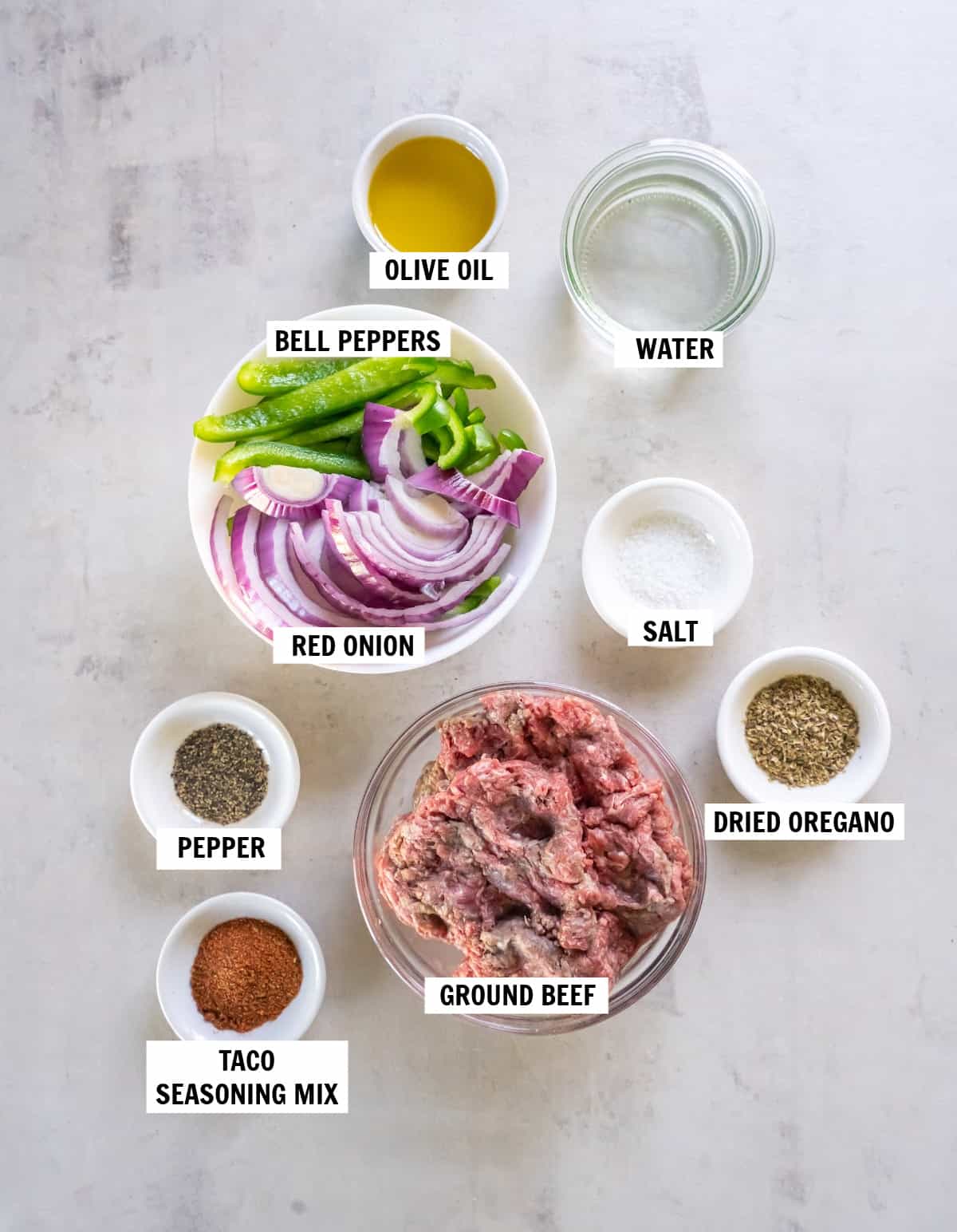 All of the ingredients for beef burrito bowls in bowls on a white countertop including olive oil, peppers and onions, salt, pepper, dried oregano, taco seasoning and ground beef.