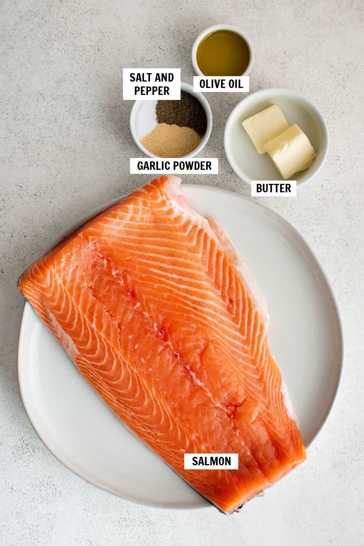 All of the ingredients for cast iron salmon on a white countertop including a salmon filet, butter, garlic powder, salt, pepper and olive oil.