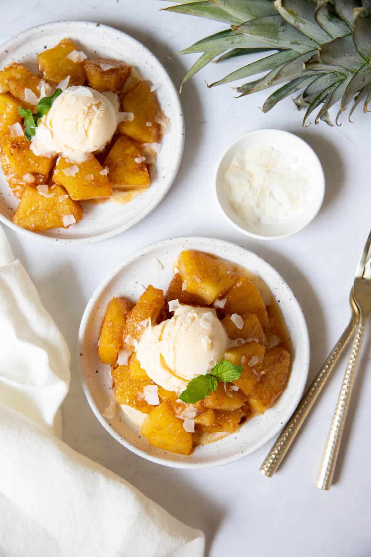 Two plates with fried pineapple and a scoop of vanilla ice cream plus coconut flakes. Forks sit to the side of the plates with a white napkin.