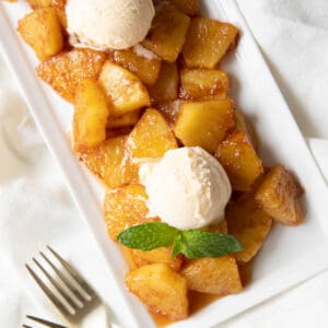 Fried pineapple served on a white serving plate with two scoops of vanilla ice cream and fresh mint leaves garnished on top. Two forks sit on the side of the plate.