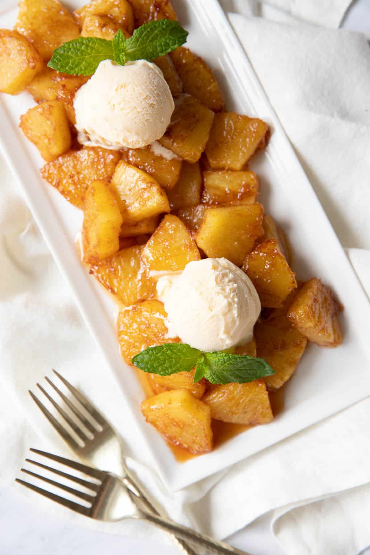 Fried pineapple served on a white serving plate with two scoops of vanilla ice cream and fresh mint leaves garnished on top. Two forks sit on the side of the plate.