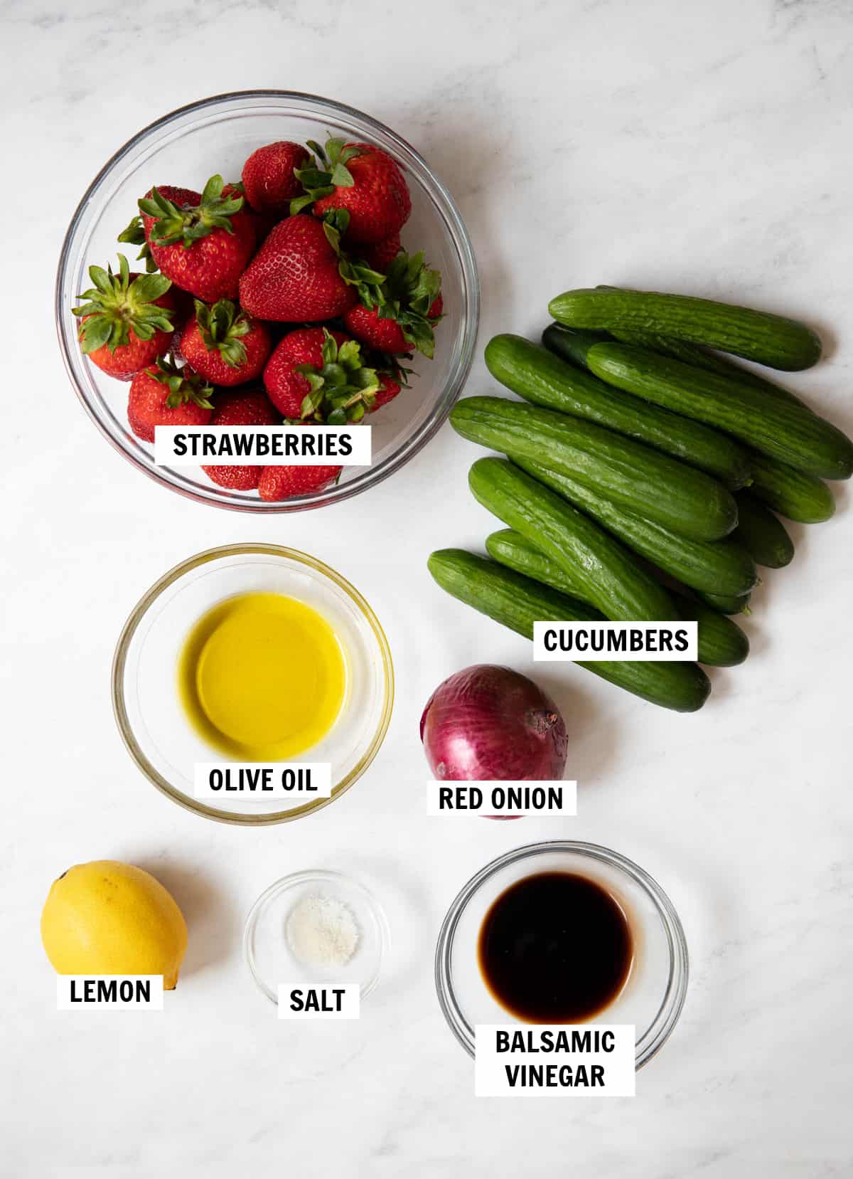 All of the ingredients for strawberry cucumber salad on a white countertop including fresh strawberries, cucumbers, red onion, lemon, olive oil, balsamic vinegar and salt.