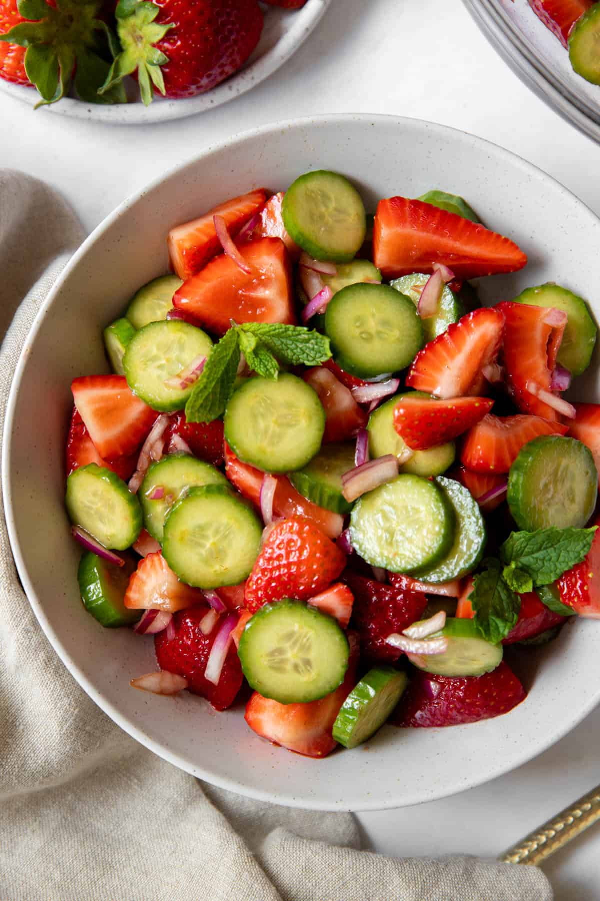 A white bowl filled with strawberry and cucumber salad that is garnished with mint. The bowl sits on a white countertop ready for serving.