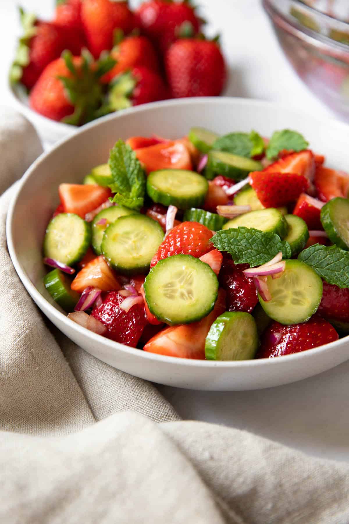 A white bowl filled with strawberry cucumber salad sits on a white tabletop with a beige napkin off to the side. A plate of fresh strawberries sits behind the bowl.