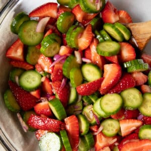 A glass bowl filled with strawberry cucumber salad ready for serving. A serving spoon sits in the bowl.