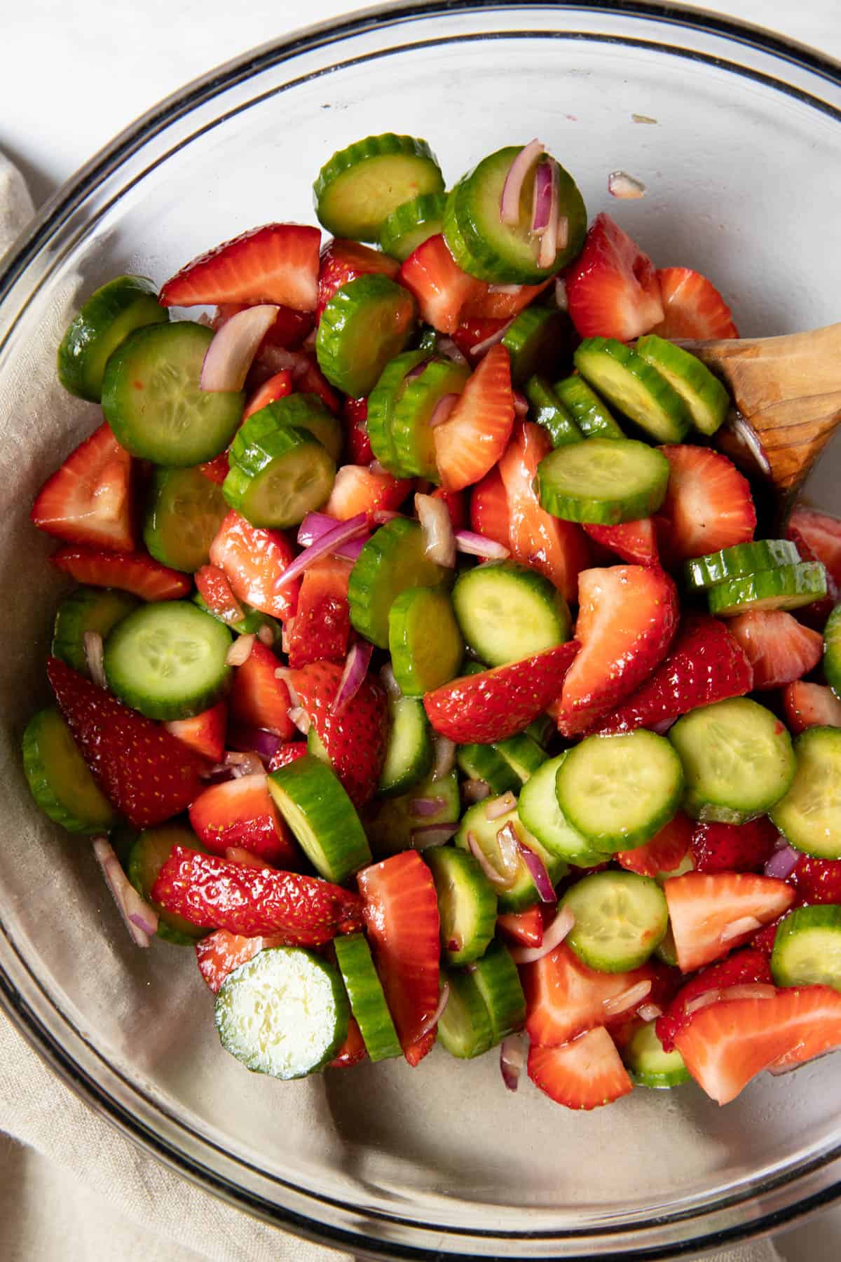 A glass bowl filled with strawberry cucumber salad ready for serving. A serving spoon sits in the bowl.