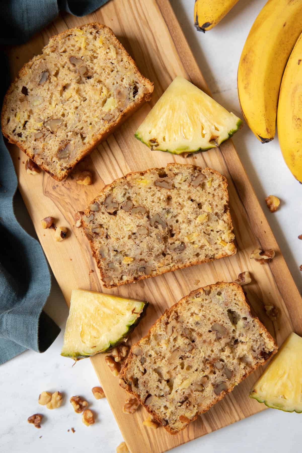 Three slices of pineapple banana bread sitting on a wooden cutting board with slices of fresh pineapple on the side.