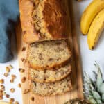 A loaf of pineapple banana bread sits on a bread board with three slices sliced off. A fresh pineapple and bananas sit next to the loaf of bread.