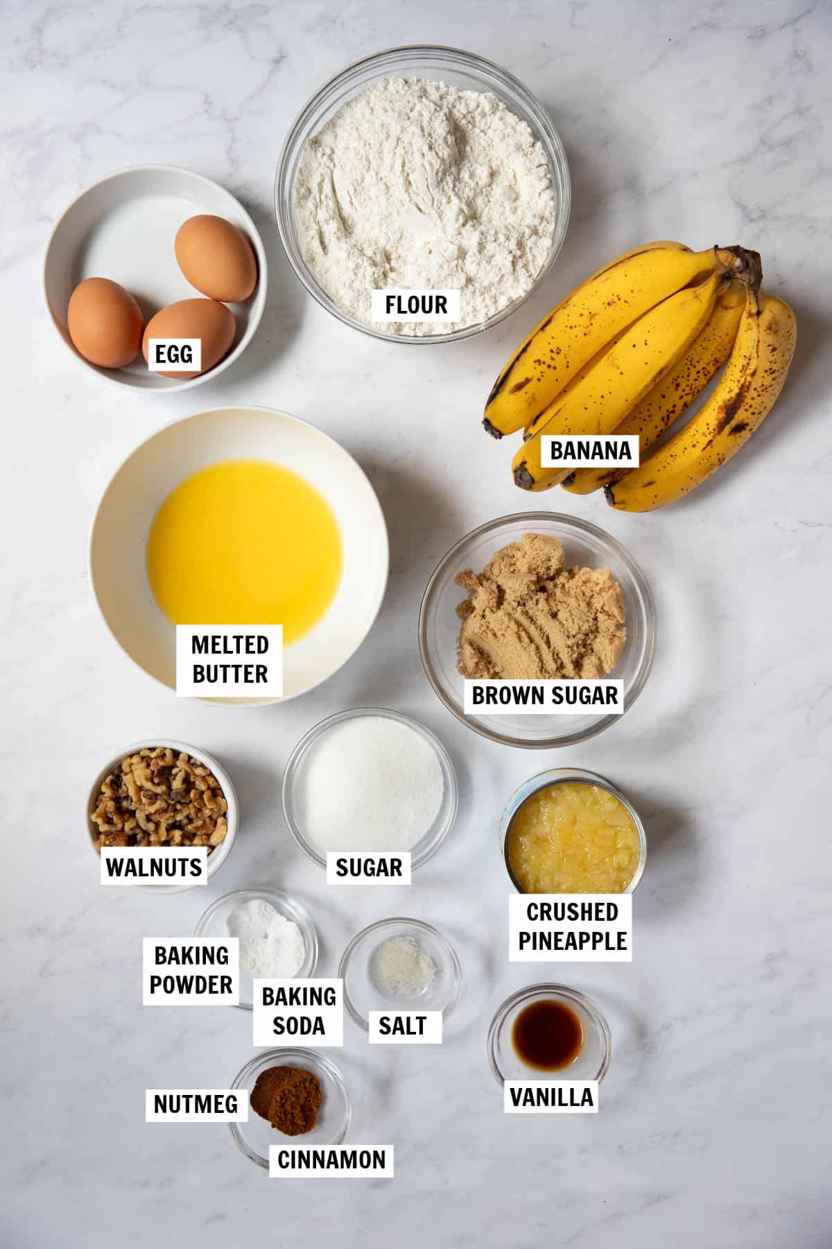 All of the ingredients for pineapple banana bread on a white countertop in bowls including flour, eggs, butter, bananas, sugar, pineapple, walnuts, baking powder and baking soda, cinnamon, nutmeg and vanilla.