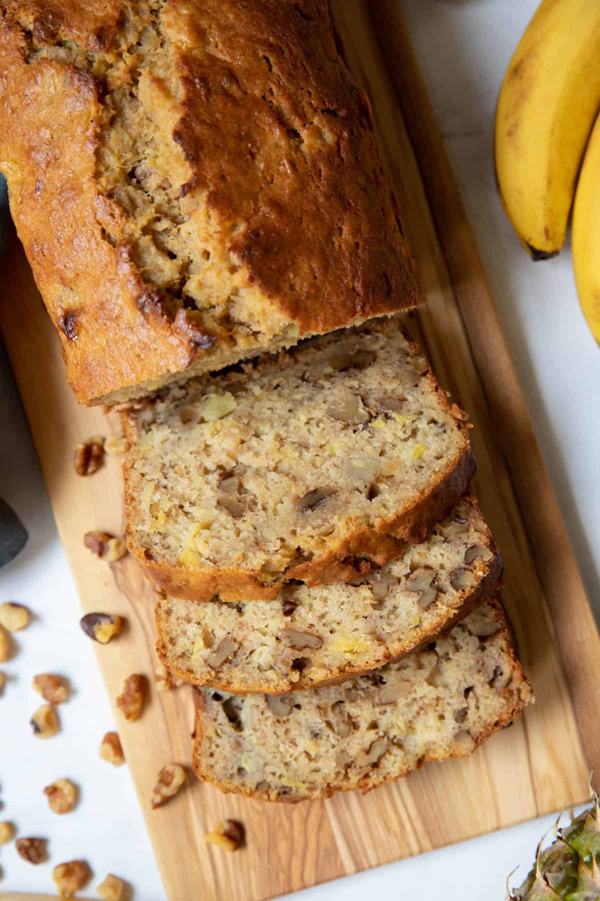 Half of a loaf of pineapple and banana bread is sliced and sitting on a wooden bread board for serving.
