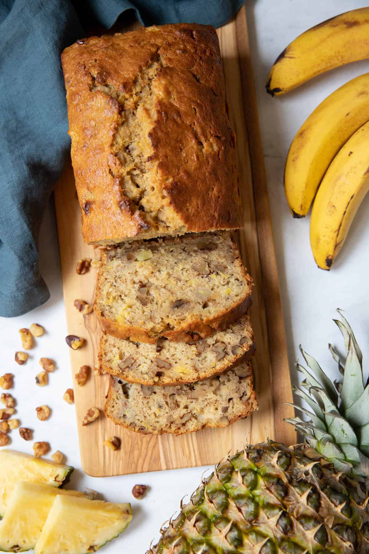 A loaf of pineapple banana bread sits on a bread board with three slices sliced off. A fresh pineapple and bananas sit next to the loaf of bread.