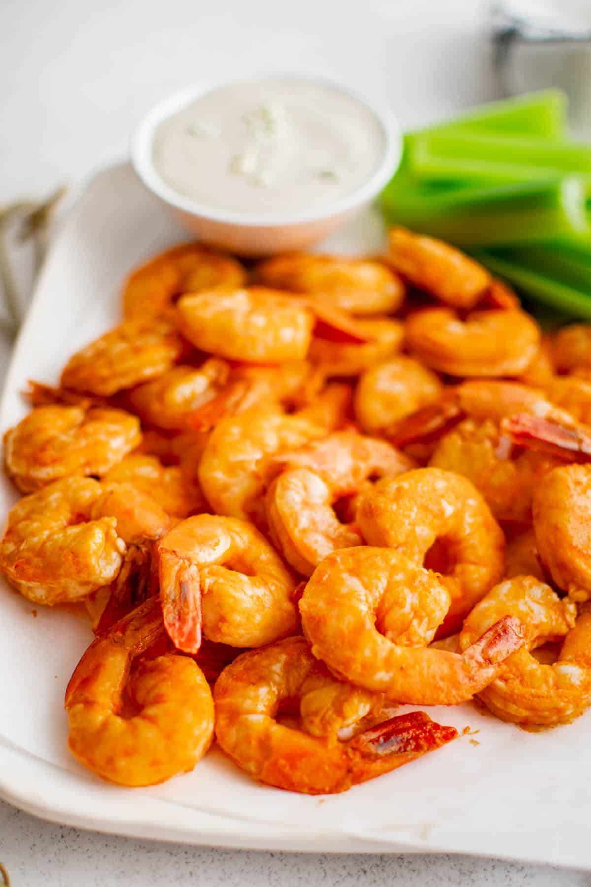 A white plate filled with buffalo shrimp and celery on the side with a small bowl of blue cheese dressing for dipping.