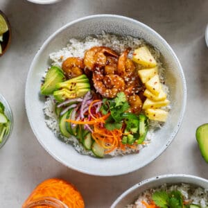 Shrimp and rice bowl in a white bowl including the cooked rice layered in the bowl with the other ingredients like the cooked shrimp, avocado, pineapple, pickled carrots and sliced cucumbers.