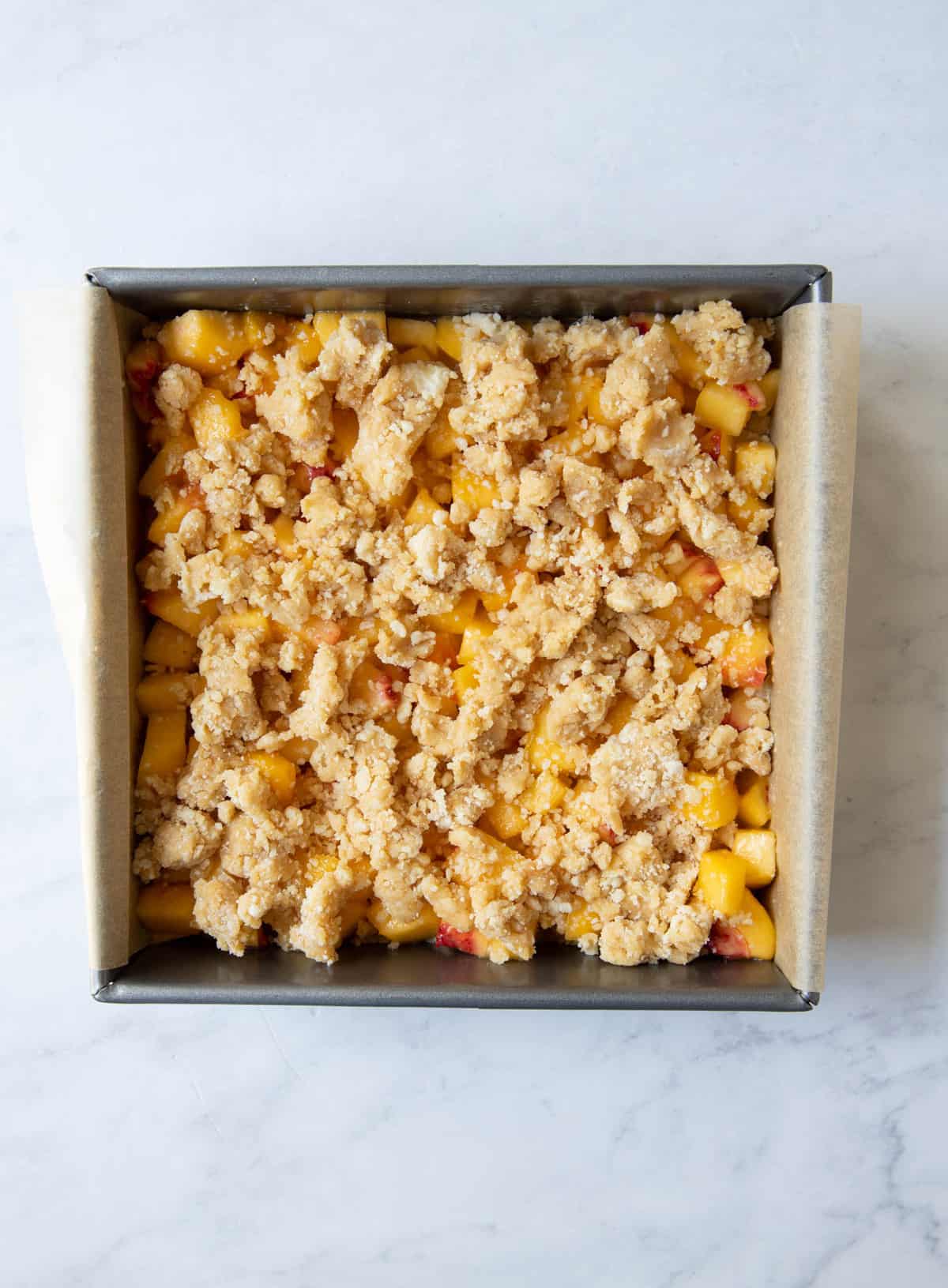 A square baking pan is layered with parchment paper, crust, peach mixture and crumb topping before baking in the oven.
