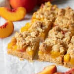 Baked peach bars sitting on white parchment paper with slices of fresh peaches off to the side.