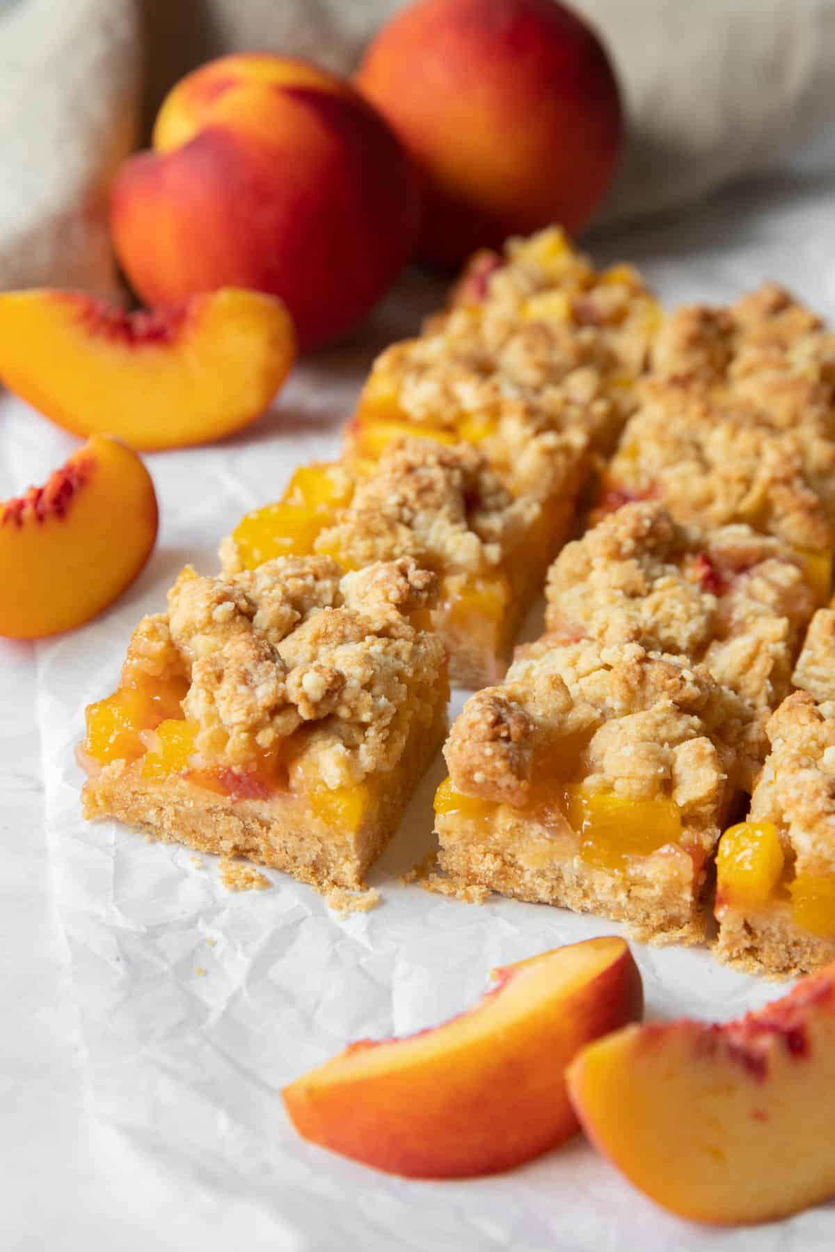 Baked peach bars sitting on white parchment paper with slices of fresh peaches off to the side.