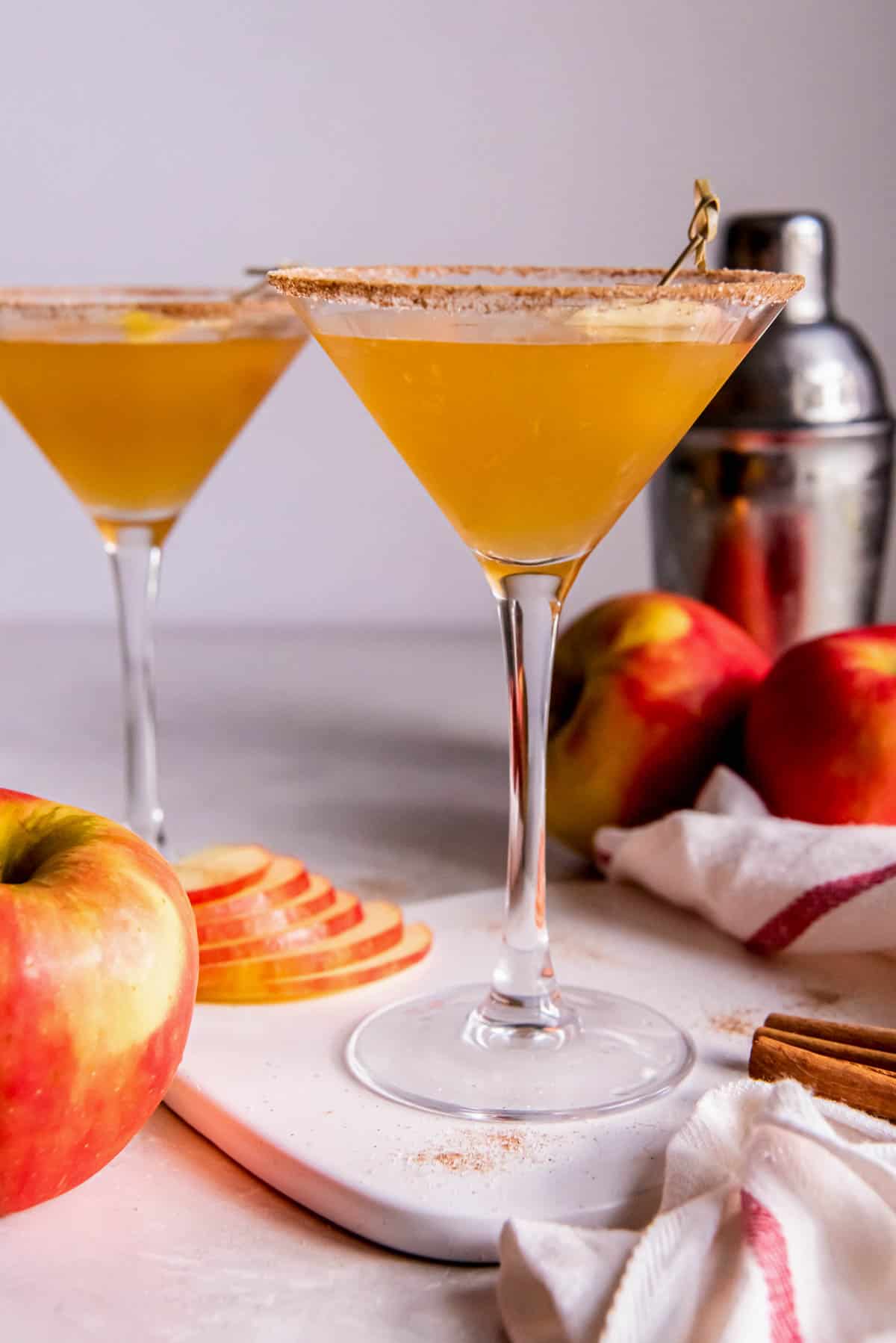 Two martini glasses with apple cider vodka martini. The glasses are rimmed with cinnamon sugar and a cocktail shaker with apples sites to the side of the drinks.