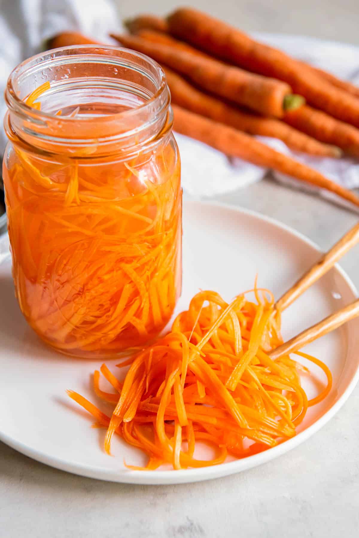 Shredded pickled carrots side on a white plate on a white countertop. The remaining jar of pickled carrots sits off to the side.