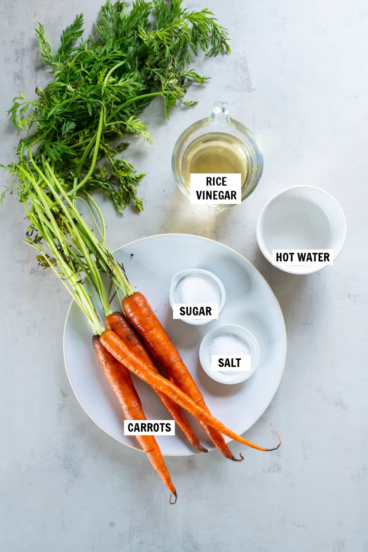 All of the ingredients for pickled carrots on a white countertop including carrots, hot water, rice vinegar, sugar and salt.