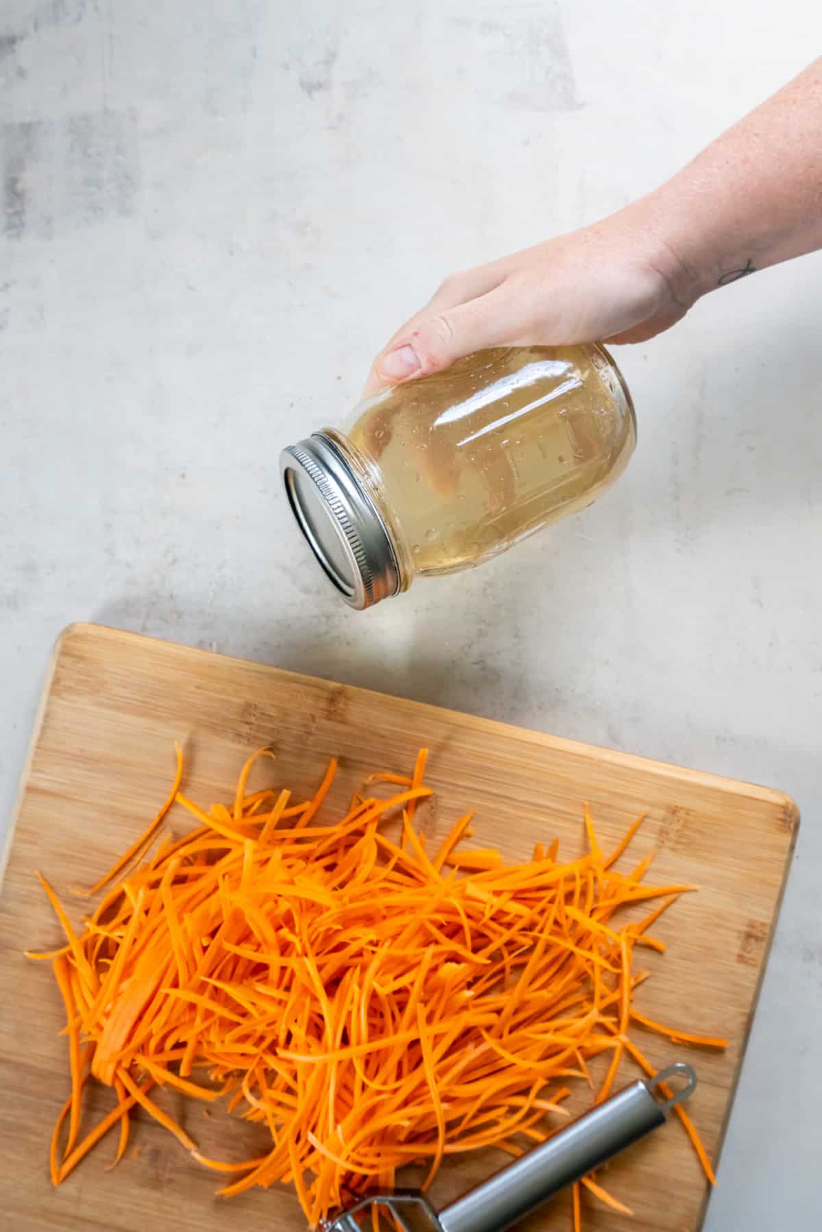 The brine for the pickled carrots in a glass jar with the shredded carrots on a wooden cutting board.
