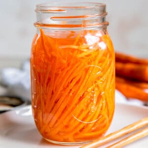 An open jar of quick picked carrots sitting on a white plate on a countertop.