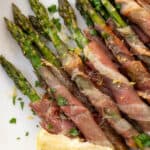 Prosciutto wrapped asparagus on a white plate garnished with lemon zest and freshly chopped parsley.