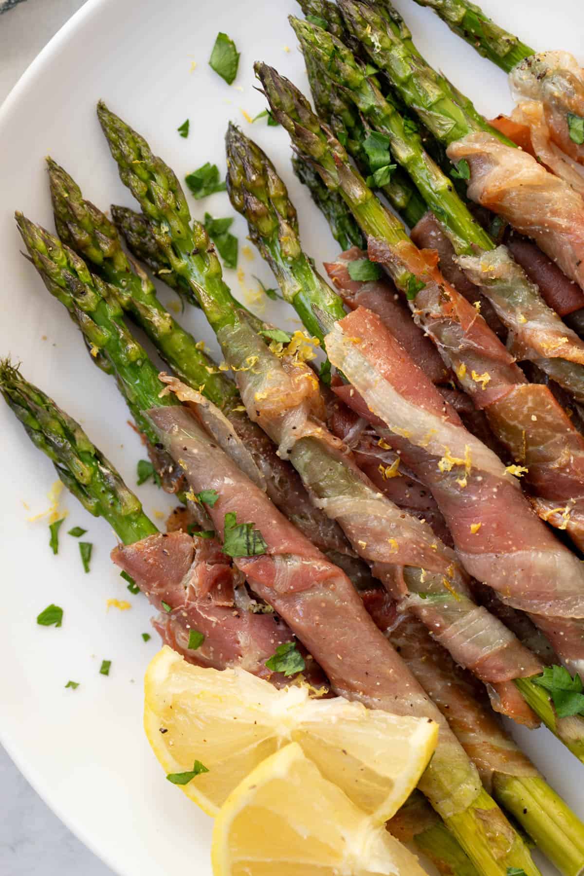 Prosciutto wrapped asparagus on a white plate garnished with lemon zest and freshly chopped parsley.