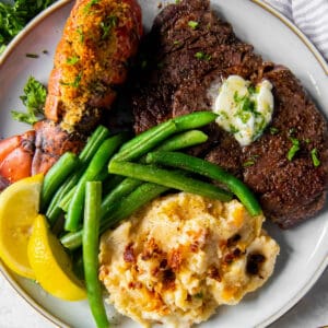 Steak and lobster served on a plate with mashed potatoes and green beans.