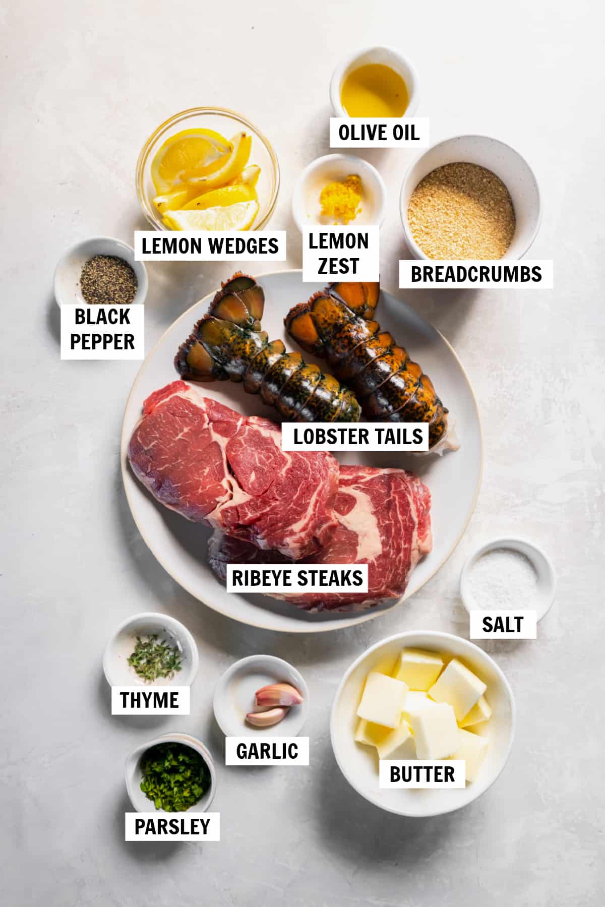 All of the ingredients for steak and lobster on a countertop in small bowls including steak, lobster tails, butter, garlic, parsley, thyme, salt and pepper, lemon zest, breadcrumbs and olive oil.