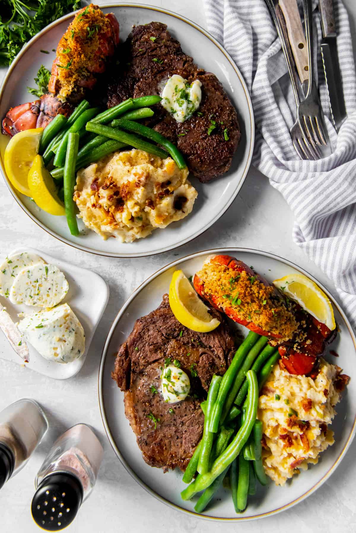Two plates with cooked lobster and steak, mashed potatoes, green beans and compound herb butter.