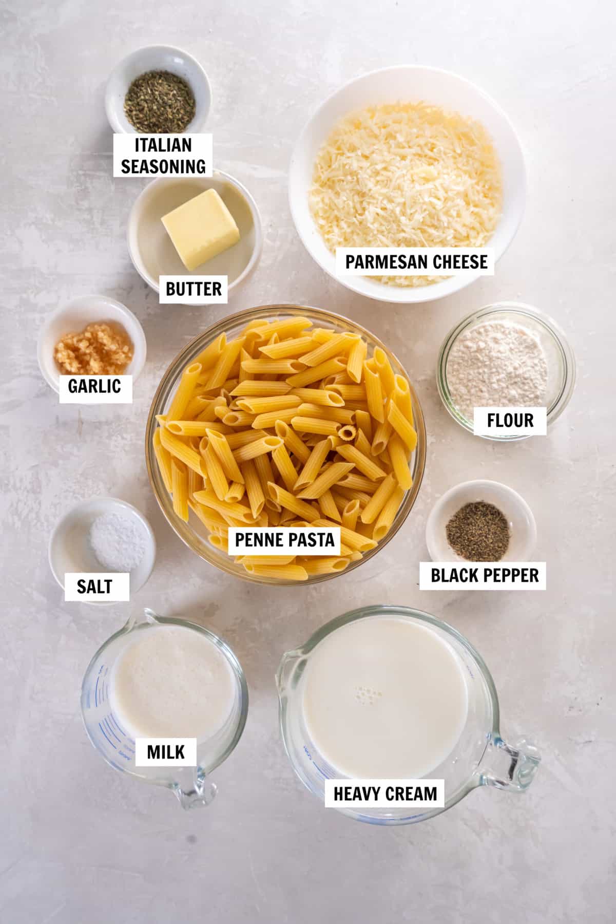 All of the ingredients you need for alfredo penne pasta in bowls on a white countertop including penne pasta, parmesan cheese, butter, milk, heavy cream, garlic, italian seasoning, salt and pepper.