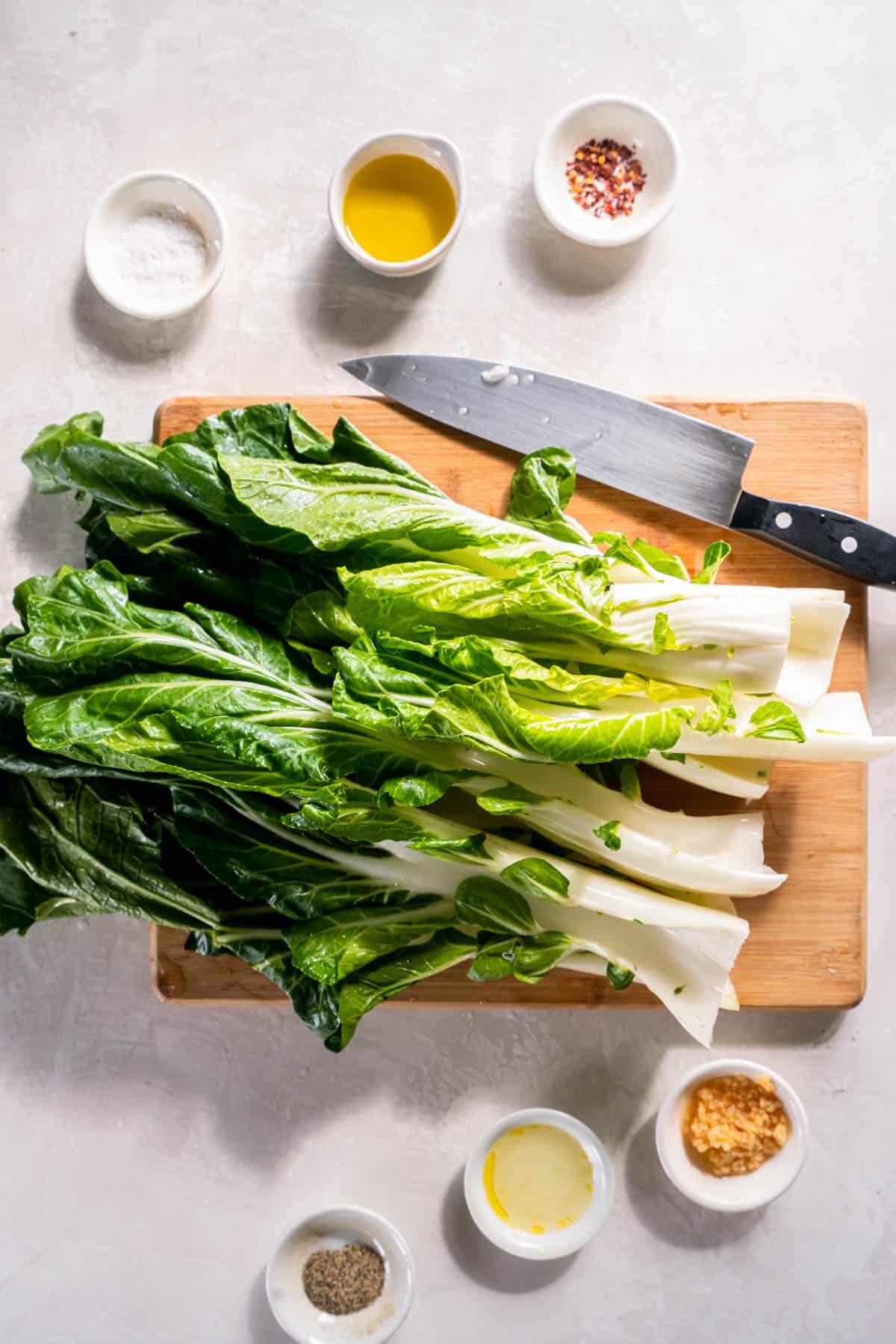 All of the ingredients for roasted bok choy on a white countertop in bowls including garlic, salt and pepper, red pepper flakes, olive oil, lemon juice and bok choy.