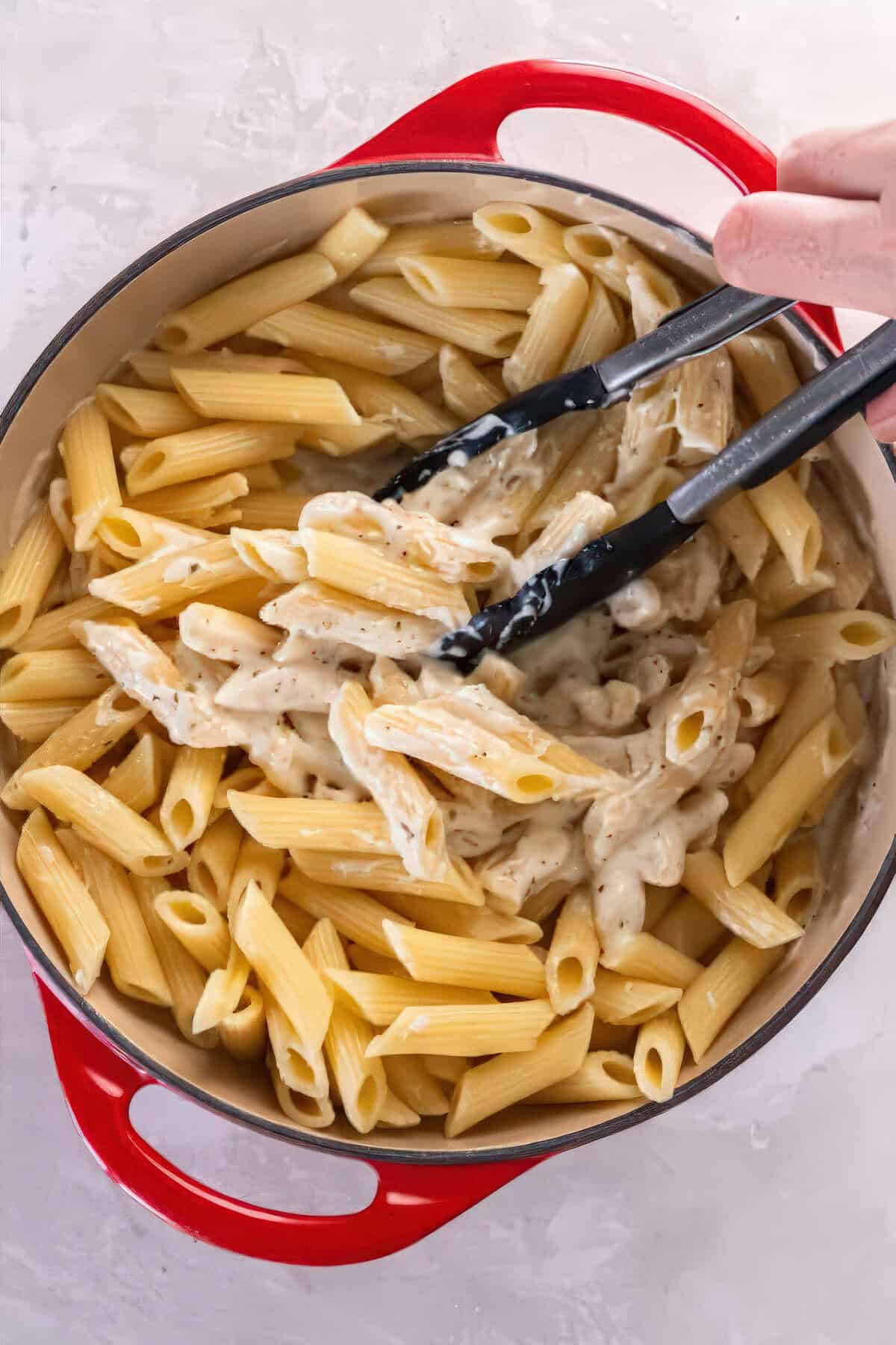 Cooked penne pasta being tossed with creamy Alfredo sauce in a red Dutch oven.