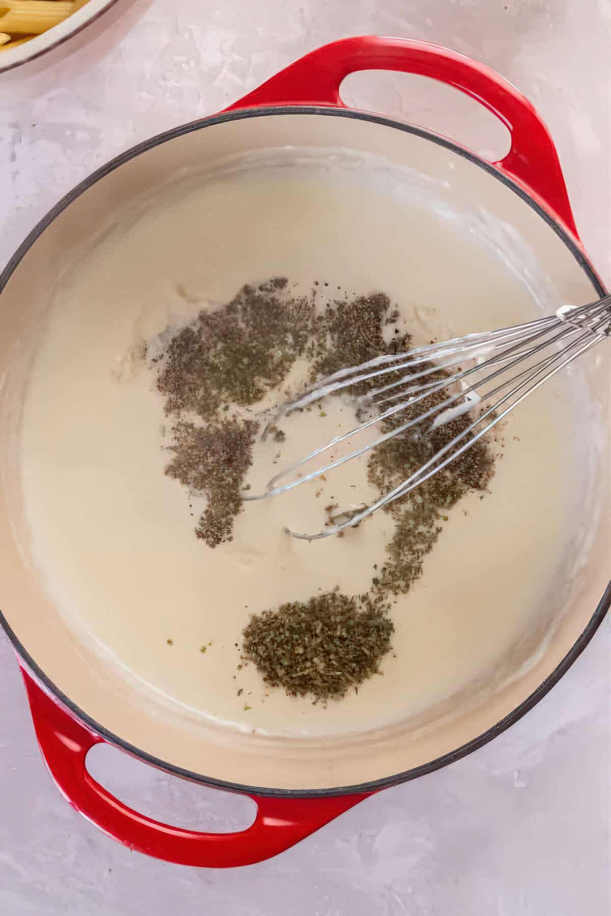 Italian seasoning and black pepper added to creamy Alfredo sauce in a red Dutch oven.