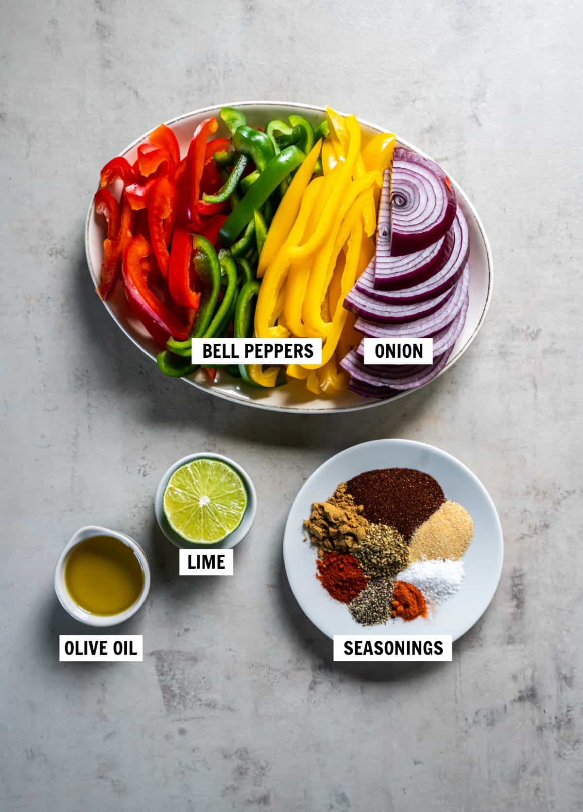All of the ingredients for veggie fajitas in plates or bowls on a gray countertop - including bell peppers, onion, olive oil, lime juice and spices.