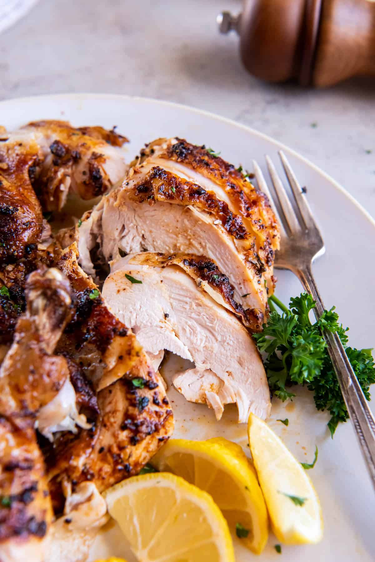 Oven roasted half chicken on a white plate sliced into small pieces for eating. A fork rests on the plate and a lemon wedge is on the side for serving.