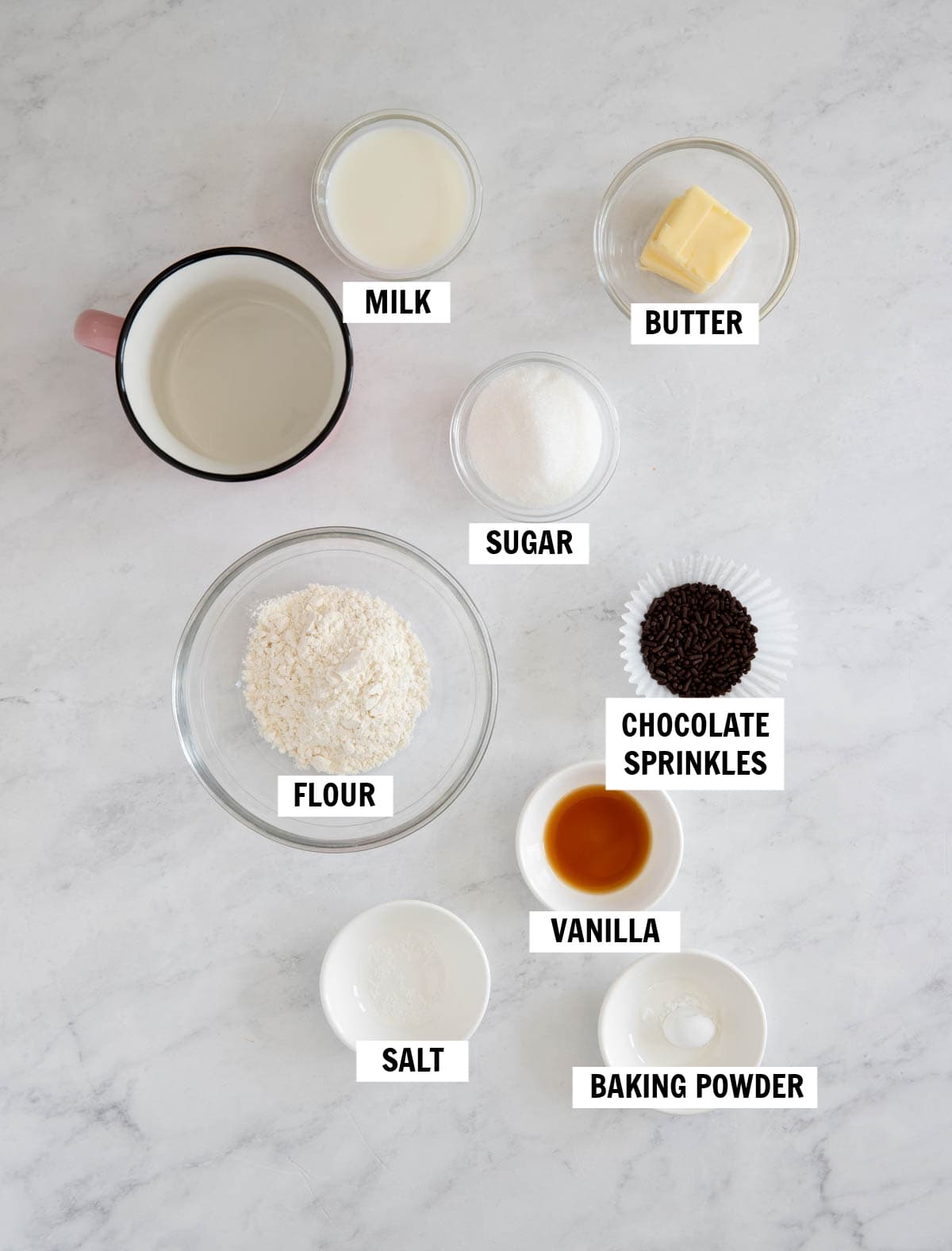 All of the ingredients for vanilla mug cake in bowls on a white countertop including flour, sugar, butter, milk, vanilla, baking powder, salt and chocolate sprinkles.