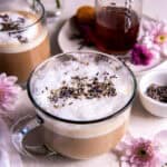 Two lavender lattes sitting on a countertop. They are garnished with fresh lavender buds.